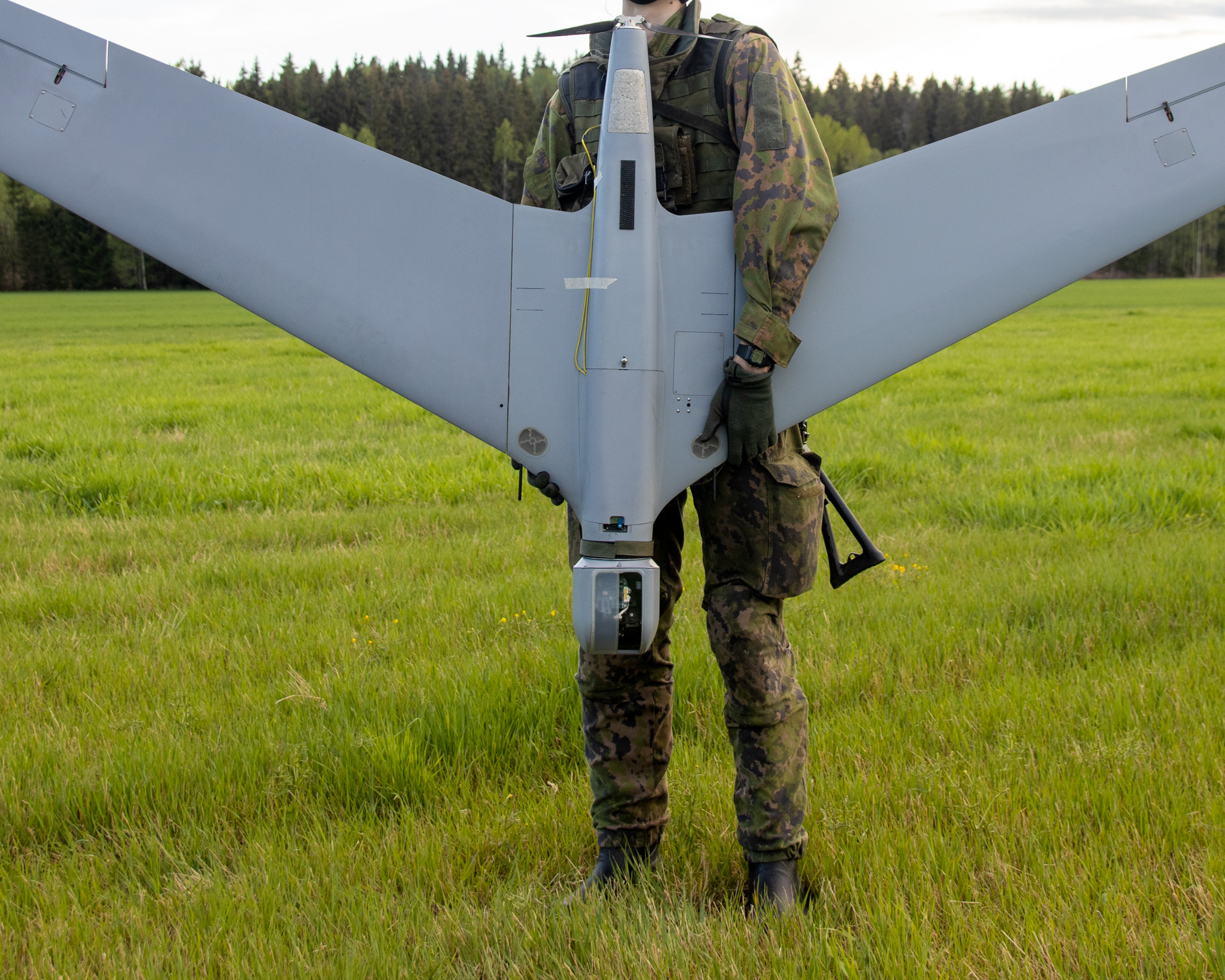 Sotilas seisoo peltoaukealla ja pitää käsissään harmaata, metallista lennokkia, taustalla metsää.