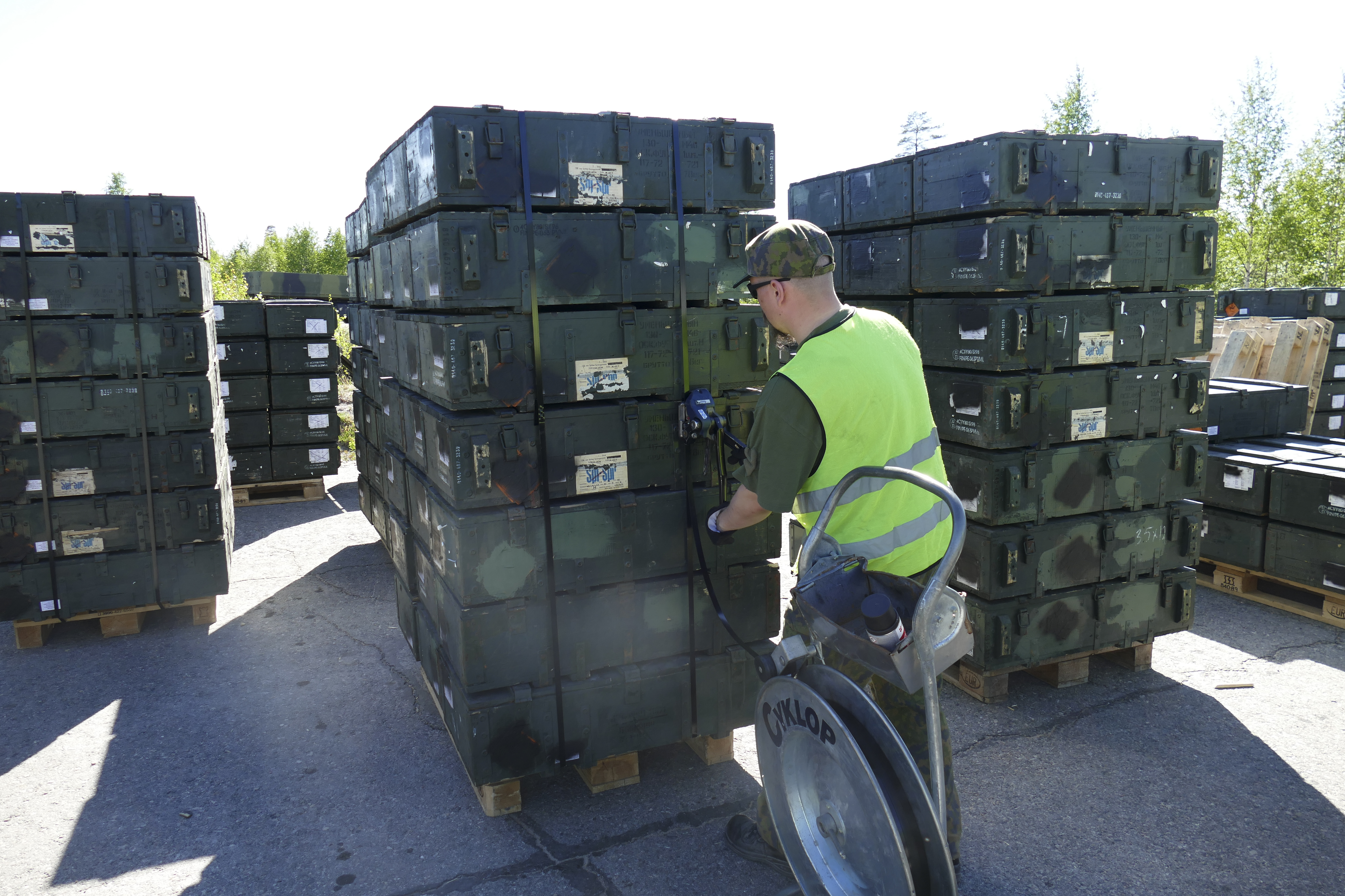 vihreitä laatikoita pinoissa kuormalavojen päällä, huomioliivinen sotilas liinoittamassa laatikkopinoa.