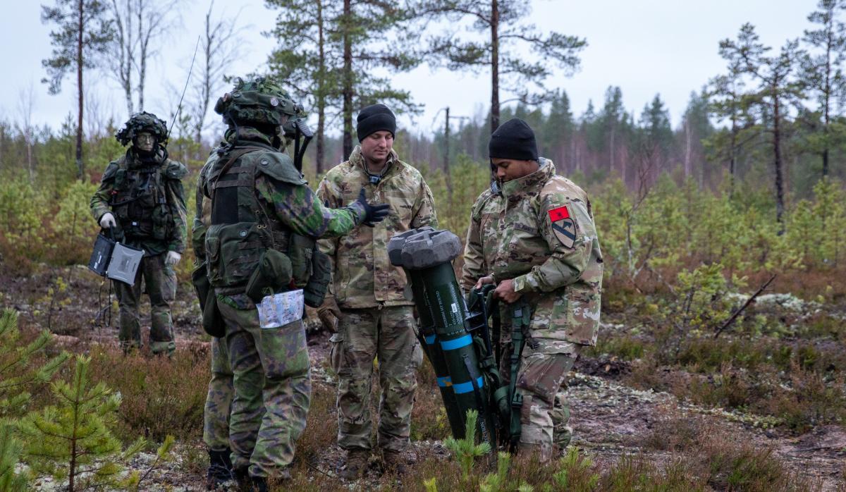 Suomalainen sotilas esittelee asettaan kolmelle yhdysvaltalaiselle sotilaalle metsämaastossa. Taustalla toinen suomalainen sotilas kävelemässä kohti.