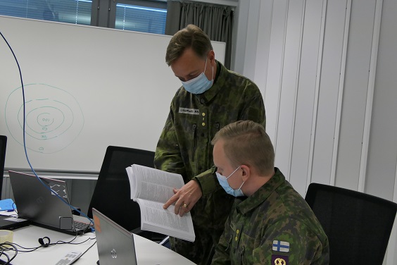 Maastopukuiset sotilaat toimistossa: seisova operaatiolakimies, reserviläinen, näyttää lakikirjasta kohtaa tietokoneen edessä istuvalle sotilaalle, yliluutnantille, henkilöstöpäällikölle.