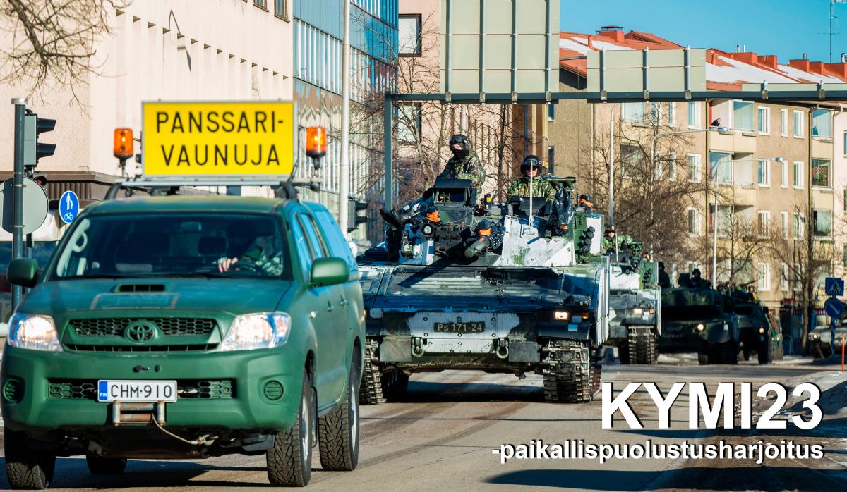 Vihreä maastoauto panssarivaunujakyltti katolla, ajaa kaupungin keskustassa ja auton perässä tulee panssarivaunuja.