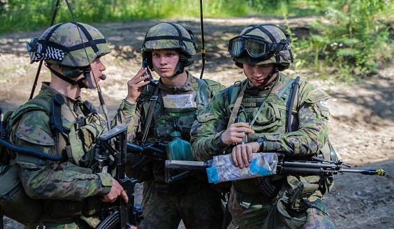 Kuvassa kolme sotilasta maastossa, yksi luurin kanssa viestimässä, kartat käsissä.