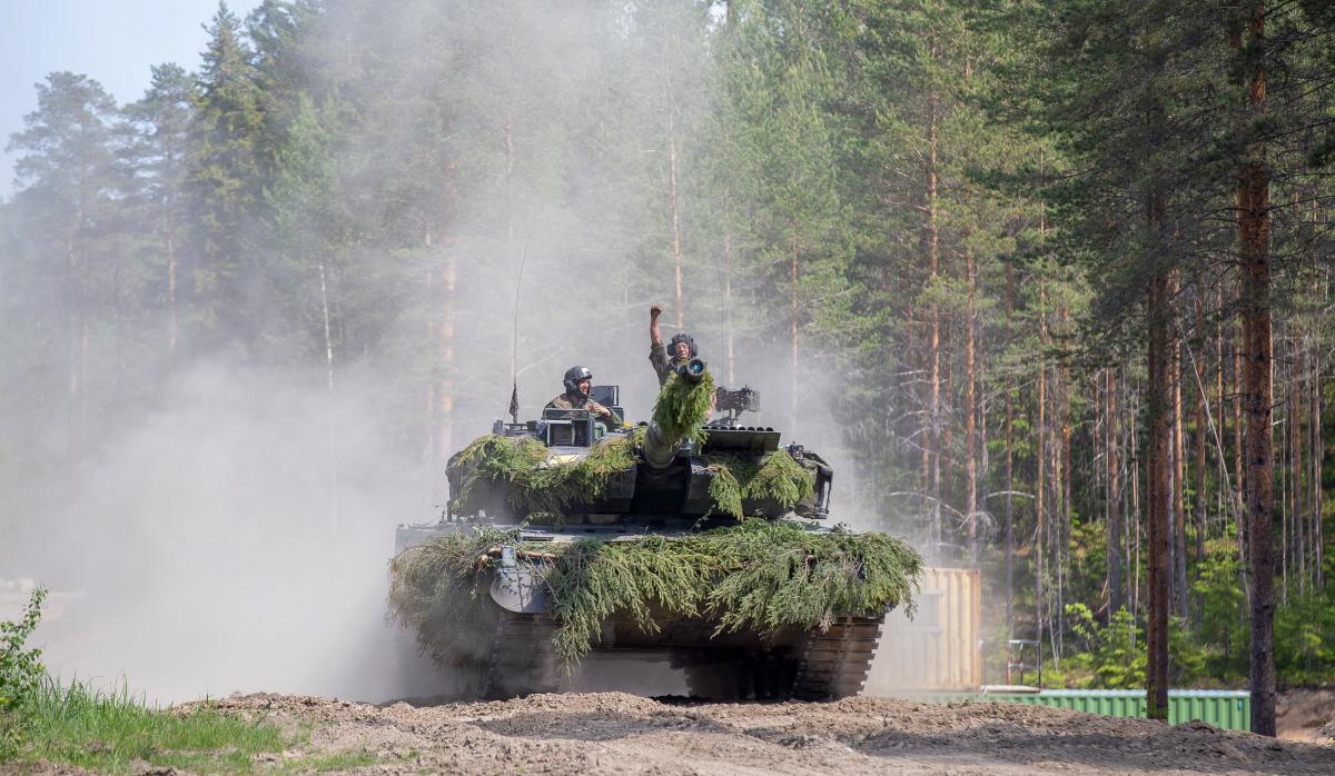 Panssarivaunu metsässä edestä kuvattuna, pölypilvi nousee takana, vaunun kuljettajalla nyrkki ilmassa huomion merkkinä.