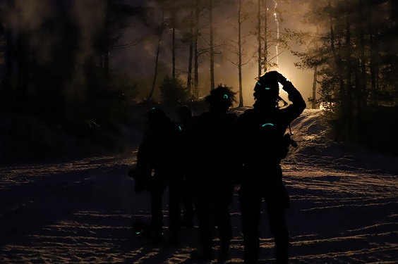 Pimeässä metsässä, talvimaisemassa tummat kypäräpäiset hahmot seisovat, joista yksi näyttää raapivat päätään.