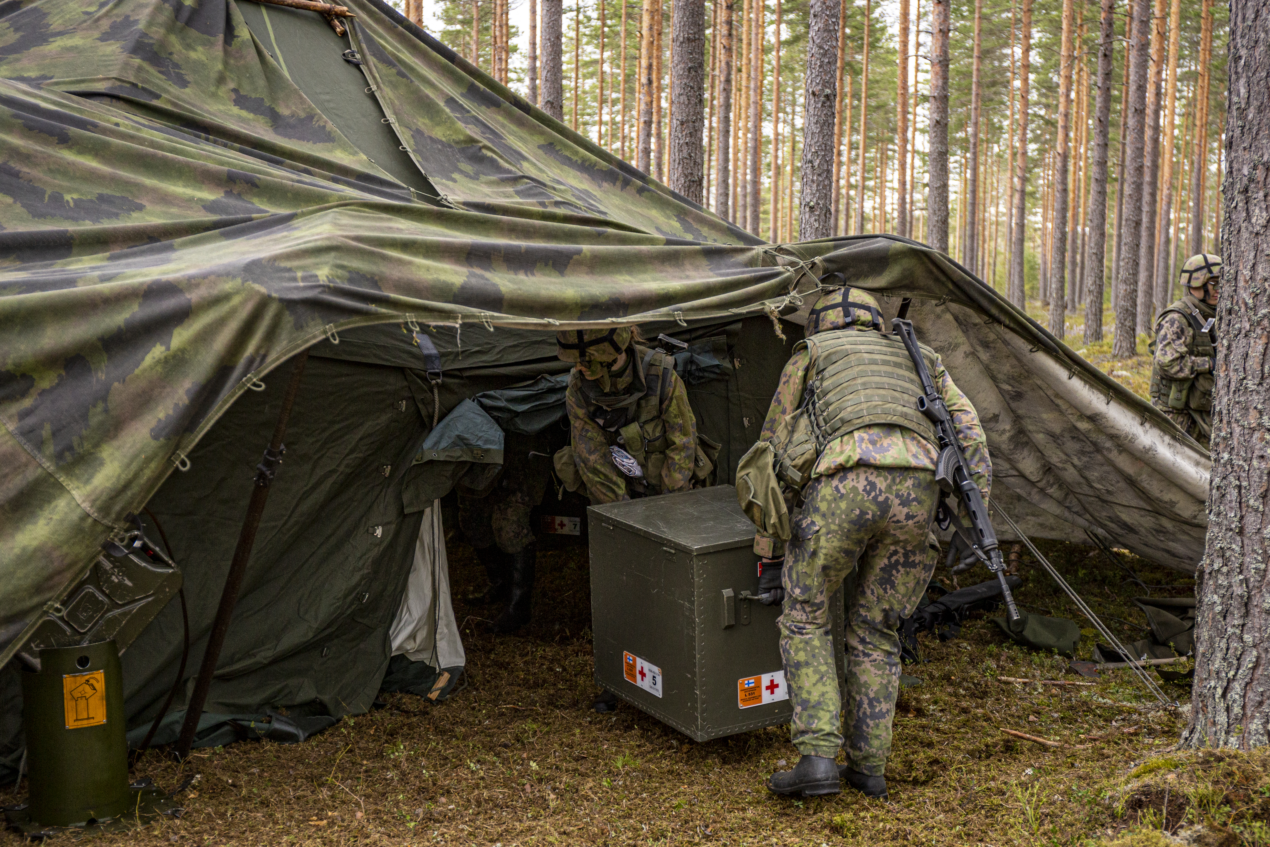 Maastokuosinen teltta metsässä, kaksi sotilasta kantaa lääkintalaatikkoa telttaan sisään.