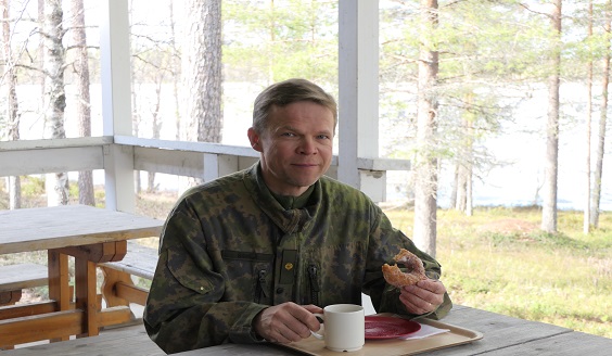 Maastopukuinen sotilas istuu pöydän ääressä luontomaisema taustalla, hänellä on kädessän sokerimunkki ja edessä kahvikuppi. Hymyilevä sotilas!