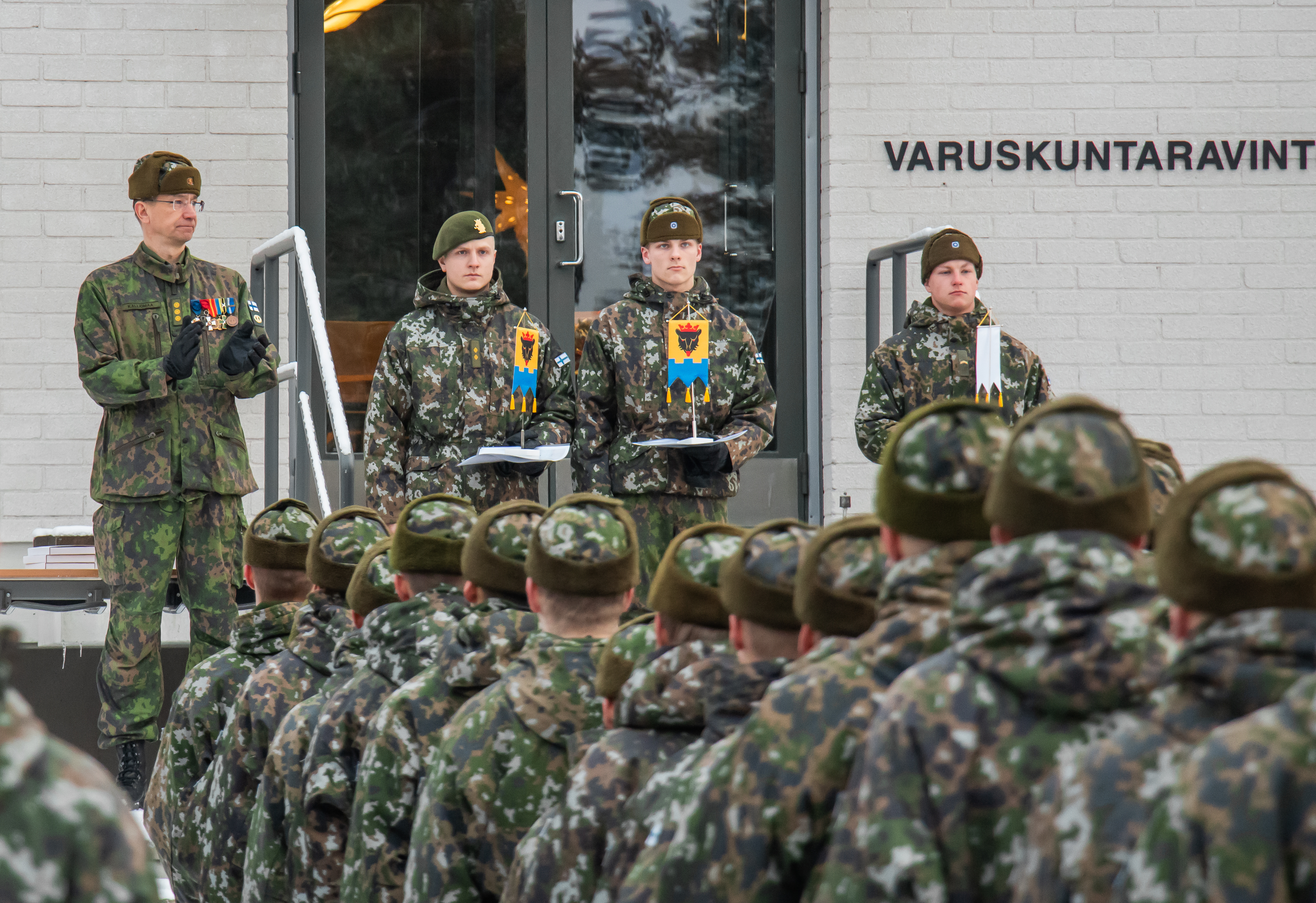 Kolme varusmiestä palkintoviirien kanssa. Heidän vieressään Eversti taputtaa ja edessä varusmiehiä seisoo muodossa.