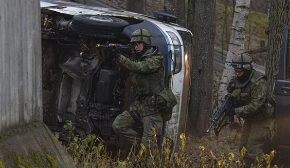 Kaksi sotilasta etenee taisteluvarustuksessa rynkkäkkökiväärien kanssa kohti betonielementtiä kyljellään olevan auton edessä