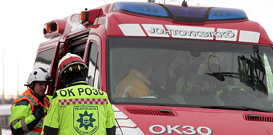 Punainen pakettiauto joss on hälytysvalot ja tuulilasin yläpuolella lukee "johtoyksikkö". Vieressä kaksi keltatakkista miestä kypärät päässä. Toisella lukee selässään "OK PO30 Pelastuslaitos Oulu-Koillismaa".