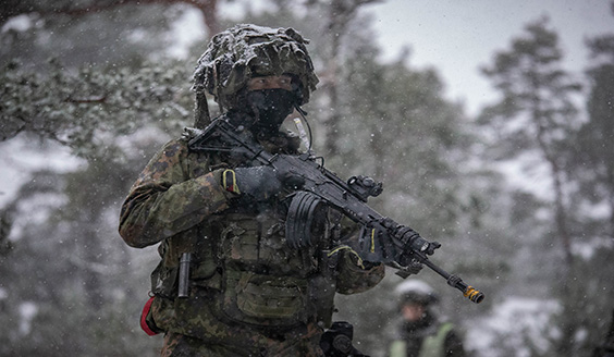 Kuvassa sotilas taisteluvarustuksessa katsoo valppaana ja pitää asetta kädessään lumisessa metsässä.