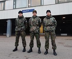 Kolme varusmeistä seisoo rakennuksen edessä. Rakennuksen seinällä on kyltti, jossa lukee Tulipatteri.