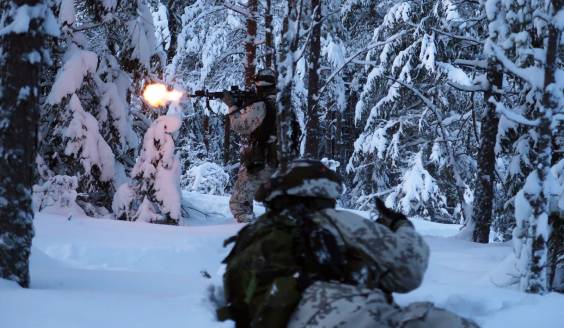 kaksi sotilasta etenevät metsässä, toinen makuuasennossa ja toinen jalkaisin samalla ampuen