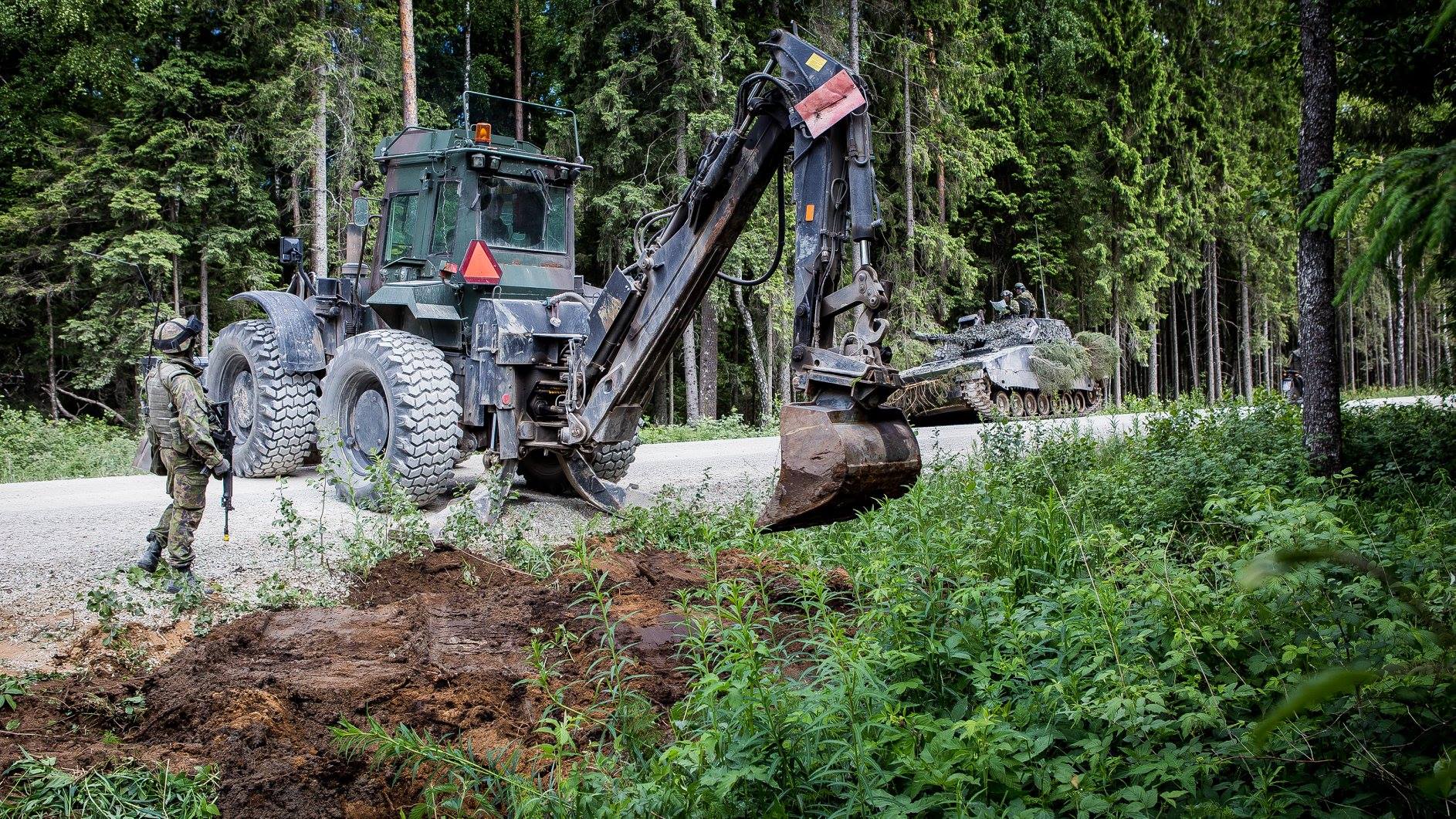 An excavator digs a small plow on a dirt road and a soldier follows next to it