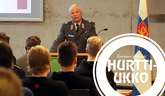 Virka-asuinen upseeri puhuu kutsunnanalaisille puhujankorokkeelta vieressään kielekkeinen valtiolippu.